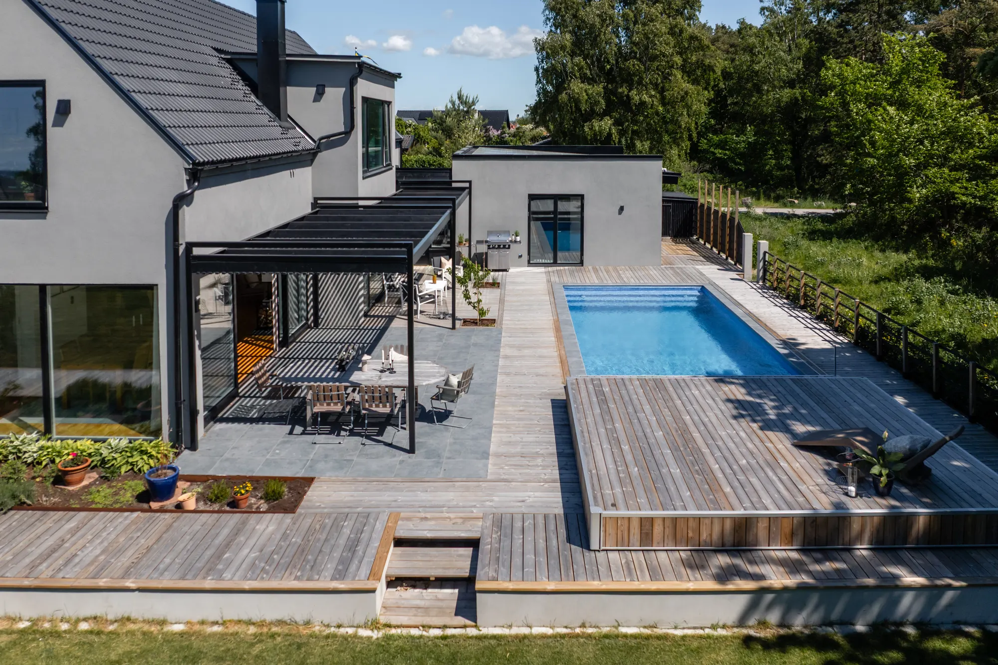 Stenhuset från ovan med terrass under skärmtak, pool och trädgård
